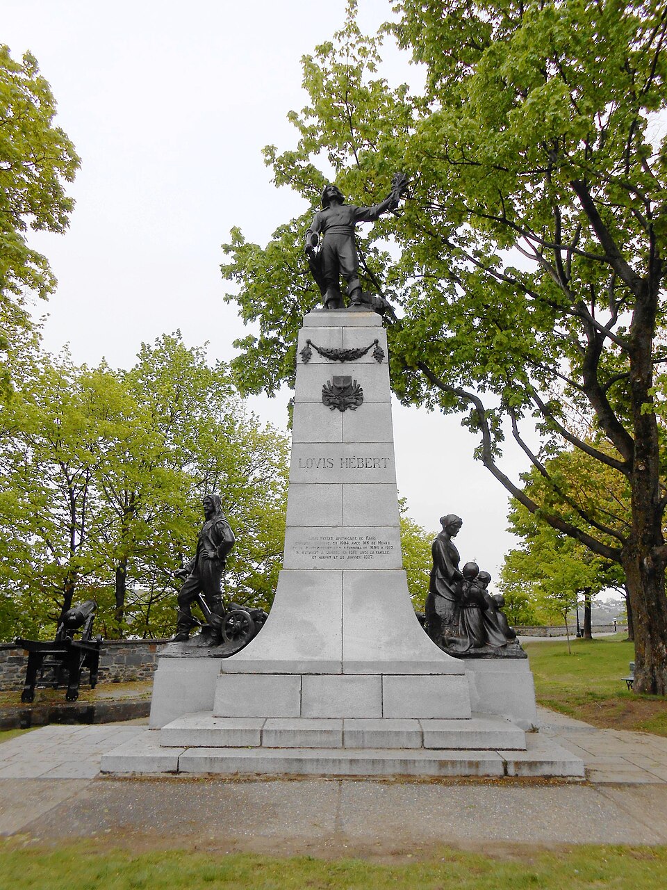 Le monument Louis-Hébert à Québec : Louis Hébert en bronze levant une gerbe de blé au sommet d'une colonne de granit, avec les statues de Guillaume Couillard et de Marie Rollet à sa base
